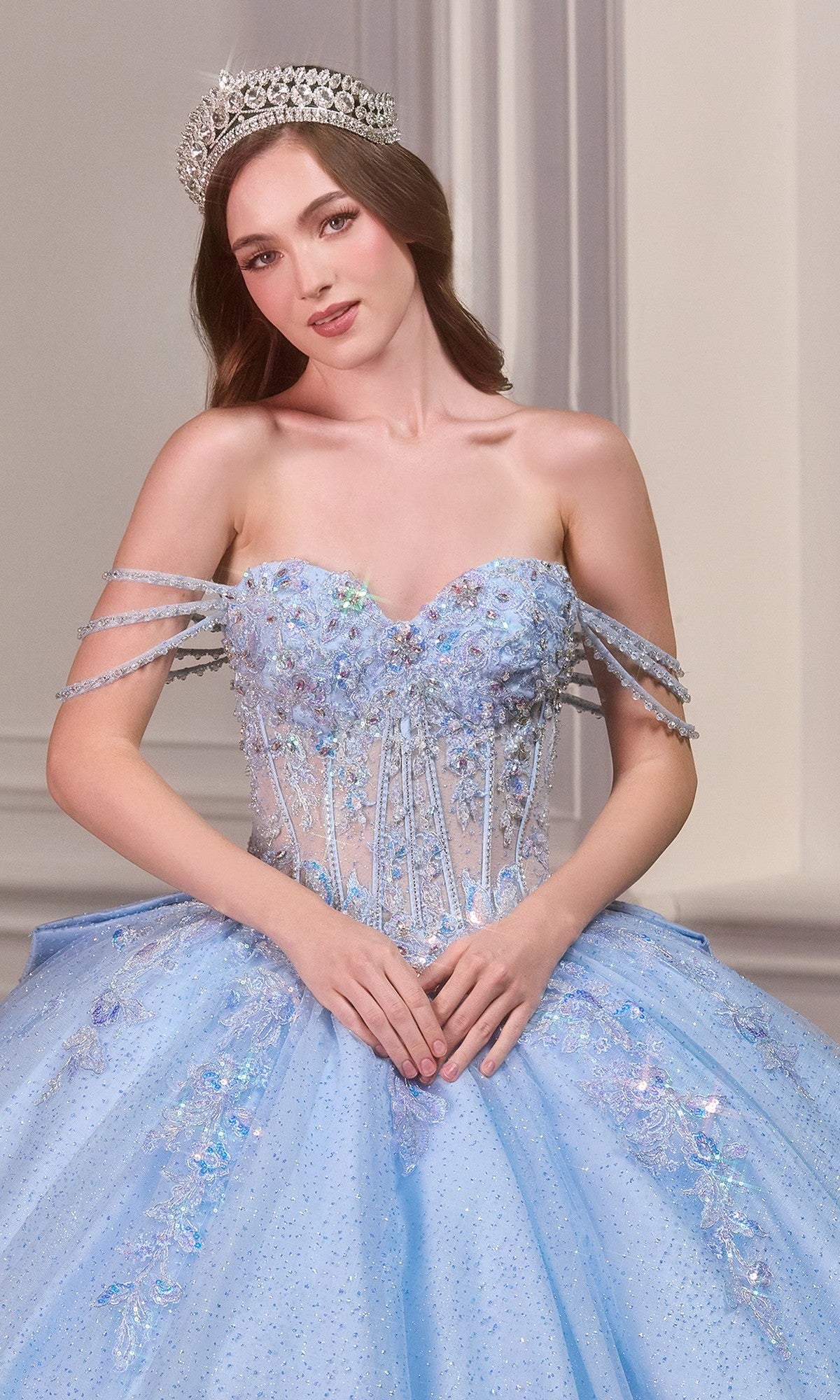 A woman in the Quinceanera Dress 15735 By Ladivine, a beaded light blue ball gown with an off-the-shoulder neckline and jeweled tiara, stands indoors facing the camera with her hands clasped.
